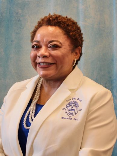 A smiling woman in a white blazer with pearls and a blue top.