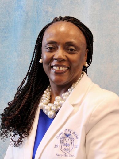 Woman with braided hair in a white sorority blazer smiling.