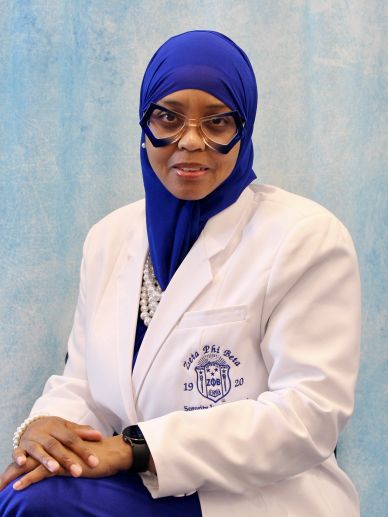 Woman in blue hijab and white blazer with glasses and pearls.