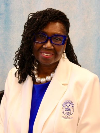 Woman in white blazer with blue glasses and pearls.