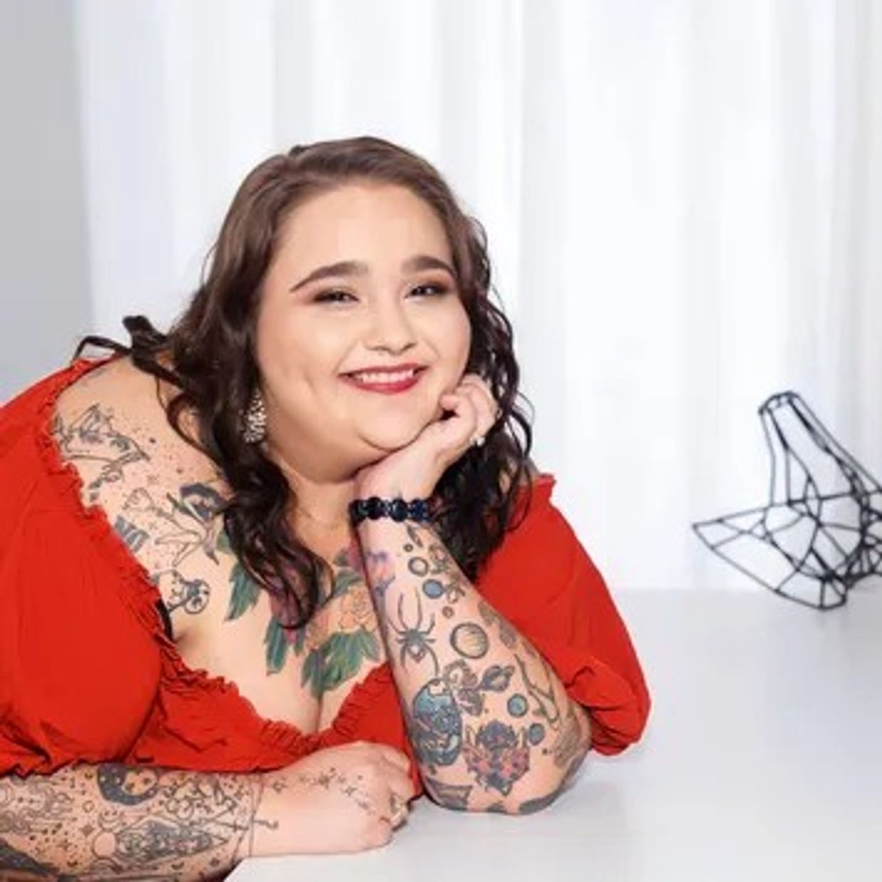 Smiling woman with tattoos, wearing a red dress, resting on a white table.