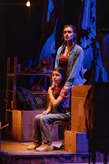 Two women in emotional moment on a dimly lit stage with boxes and shelves.