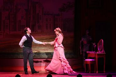 Actors dancing on stage in period costumes with a castle backdrop.