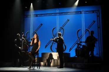 A female singer performs with a band on a stage with blue violin-themed backdrop.