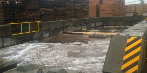 Industrial site with stacks of lumber and a containment area filled with liquid.