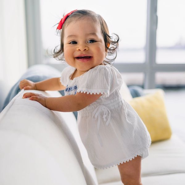 Infant girl in white dress with red bow stands by and holds onto a white sofa.