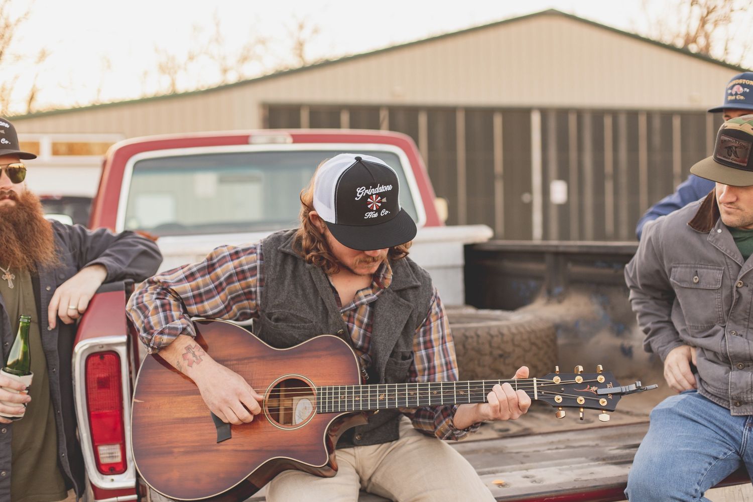 A man playing guitar while sitting in the back of a pick-up truck with three men watching.