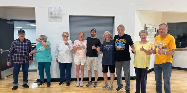 A diverse group of people standing in a gym, smiling and giving thumbs up.