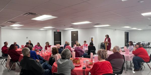 A group gathered for a community event in a hall with pink tablecloths and decorations.