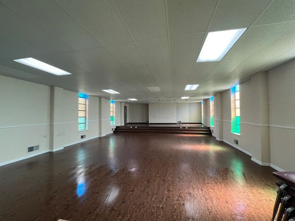 Empty room with wooden floor, stained glass windows, and a raised stage area.