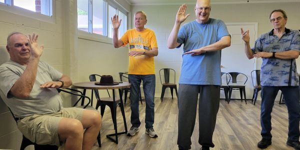 Four men practicing Tai Chi in a bright room with wooden floors.