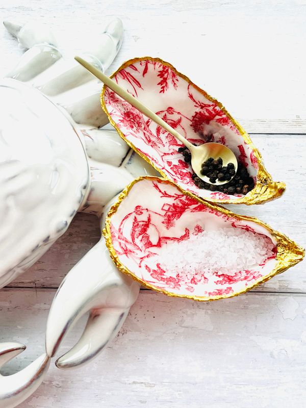 Decorative shell-shaped dishes with gold edges holding salt and pepper beside a white ceramic lobster.