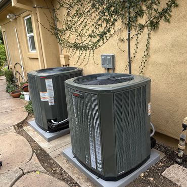 Two outdoor air conditioning units installed beside a beige house wall.