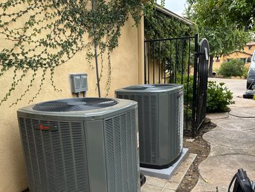 Two outdoor air conditioning units installed against a house wall with a pathway beside them.