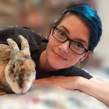 Person with blue hair and glasses lying next to a rabbit.