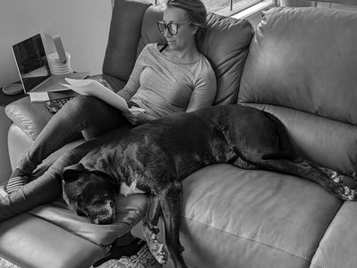 Sarah Schmitt sitting on a couch copyediting a document while her dog lies on her.