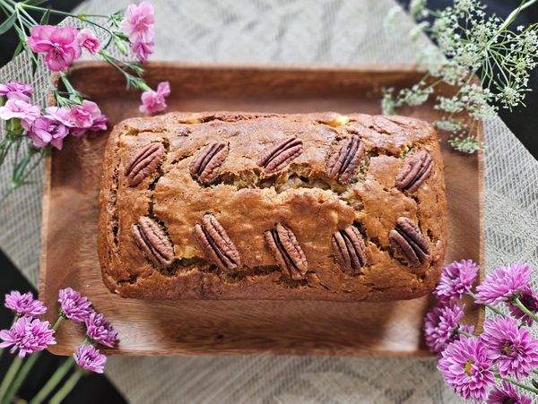 Freshly baked pecan-topped loaf cake on a wooden board surrounded by flowers.
