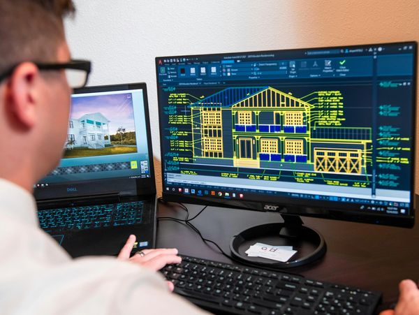 Person designing a house using CAD software on dual monitors.