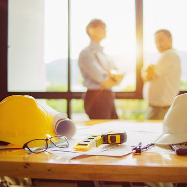 Construction plans and safety gear on a desk with two engineers in background.