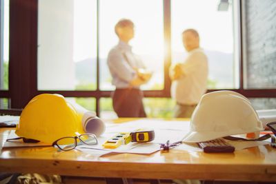 Construction helmets and tools on a desk with two people discussing in the background.
