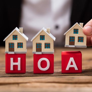 Miniature houses on blocks spelling HOA, one held by a hand.