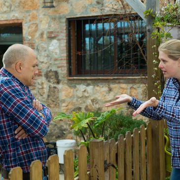 Two people arguing over a wooden fence in a garden area.