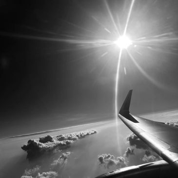 Black and white photo of an airplane wing with the sun shining above clouds.