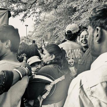 A crowded outdoor scene with people, including a woman carrying a baby in a carrier.
