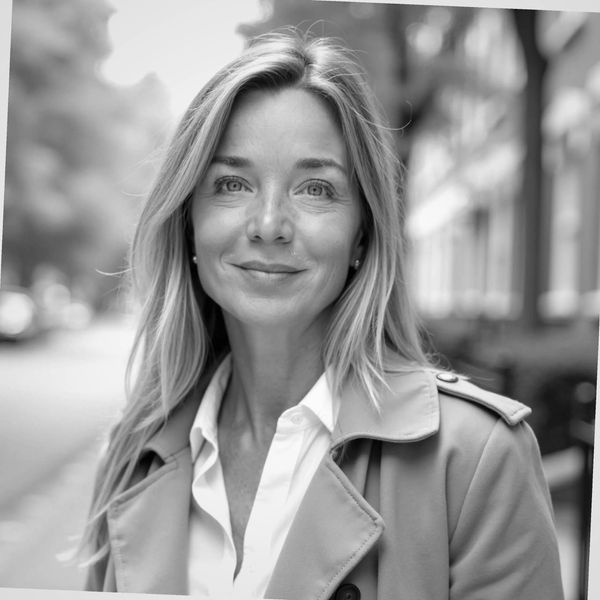 Confident woman with blonde hair wearing a tan coat and white shirt outdoors.