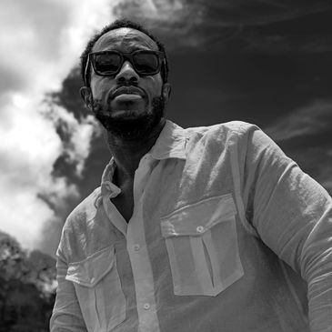 Portrait of a man in sunglasses and casual shirt against a cloudy sky.