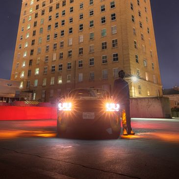 Car with headlights on parked at night near a tall building.