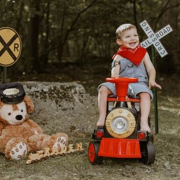 A smiling toddler in overalls rides a toy train outdoors with a teddy bear and railroad signs nearby.