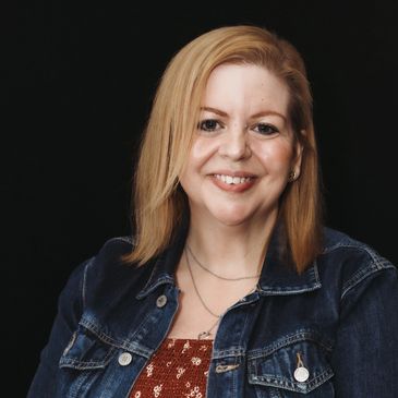 Smiling woman with blonde hair wearing a denim jacket against a black background.