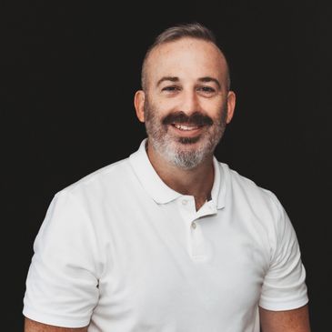 Smiling man with a beard wearing a white polo shirt against a black background.