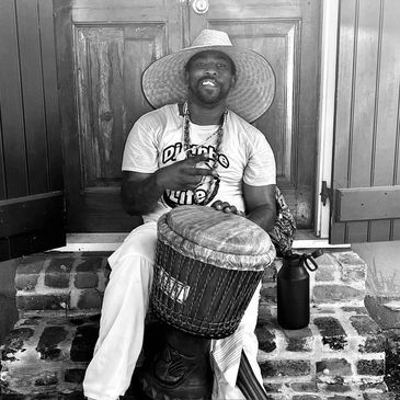 Man playing a drum outdoors, wearing a large hat.