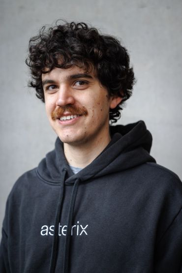 Young man with curly hair and mustache in a black hoodie.