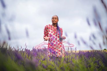 Woman in floral dress stands in lavender field holding a pink wire hanger.