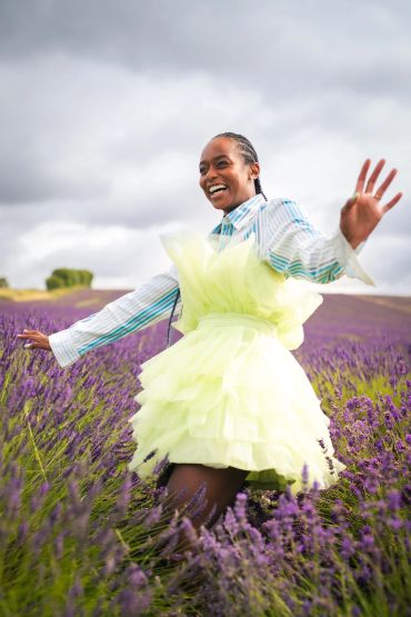 Smiling woman in a yellow dress joyfully stands in a lavender field under a cloudy sky.