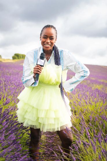 A smiling woman in a bright dress stands in a lavender field holding a microphone.