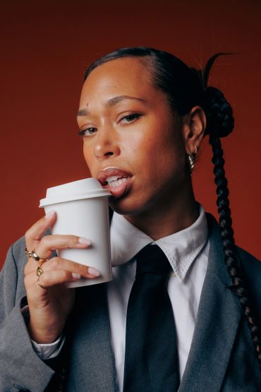 Person with braided hair sipping coffee in a suit.