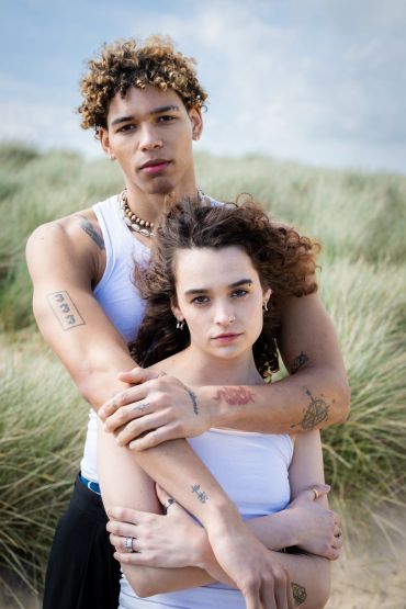 Couple embracing outdoors with a backdrop of tall grass under a cloudy sky.