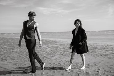 Two stylish individuals walk barefoot on a beach in a black and white photo.