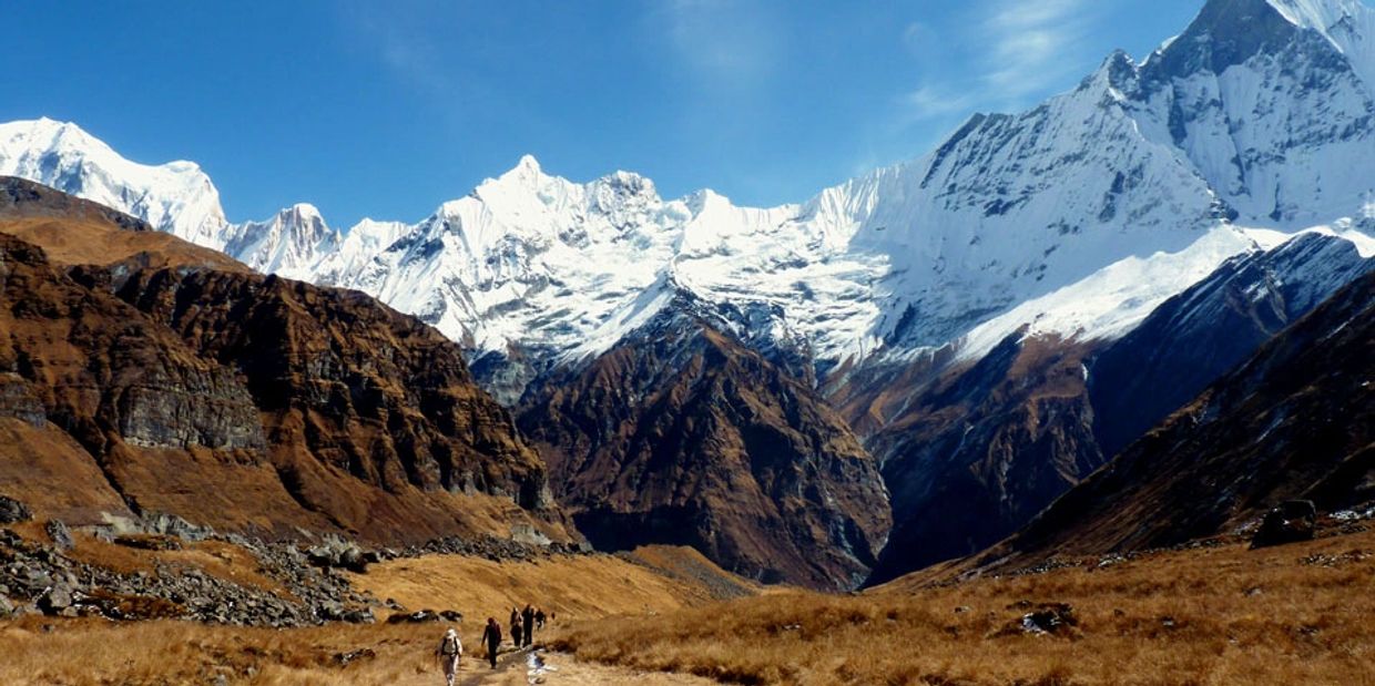 Hikers trekking through a valley surrounded by snow-capped mountains under a clear blue sky.