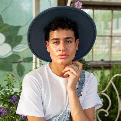 Person with curly hair wearing a wide-brimmed hat and overalls in a garden.