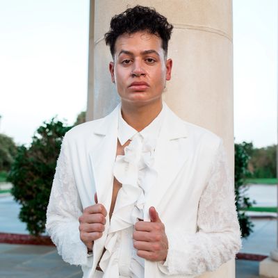 Person in a white ruffled shirt and blazer posing against a pillar.