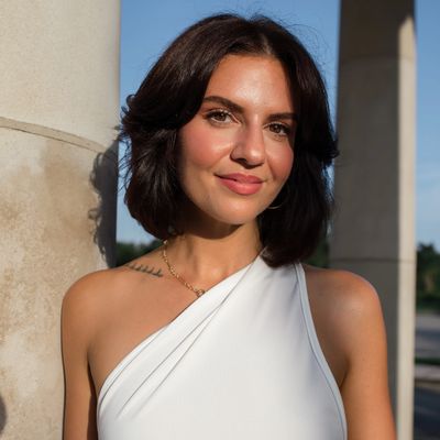 Woman with dark hair in white one-shoulder top smiling.