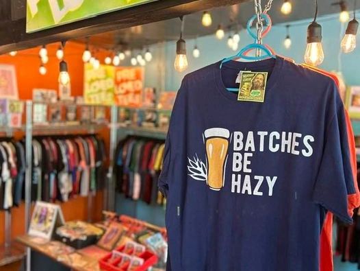 A navy blue t-shirt with a beer-themed slogan hangs in a colorful store.