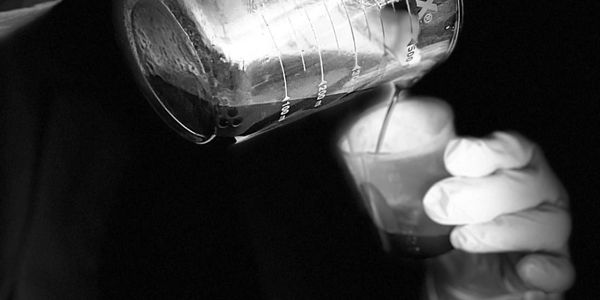 Gloved hands pour liquid from a Pyrex measuring cup into a small container.