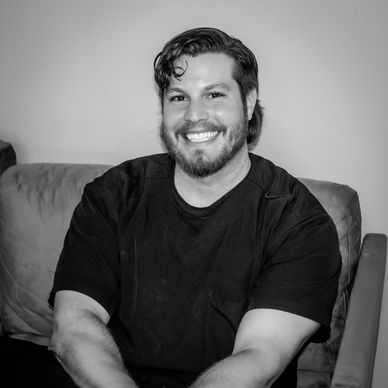 A smiling man with a beard sitting on a couch in a black shirt.