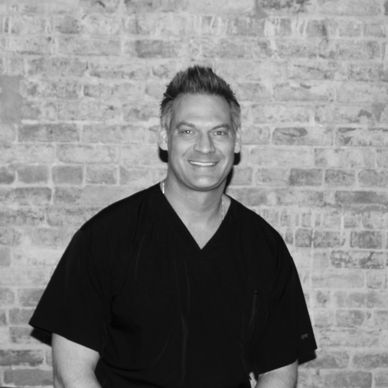Man in black scrubs smiling against a brick wall.